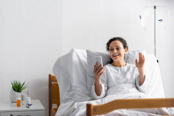 femme afro-américaine souriante agitant la main lors d'un appel vidéo sur smartphone à l'hôpital - Photo, image