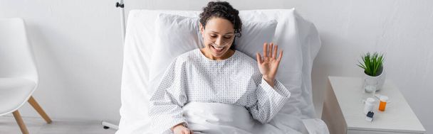 весела афроамериканка махає рукою, сидячи в лікарняному ліжку, банер
 - Фото, зображення