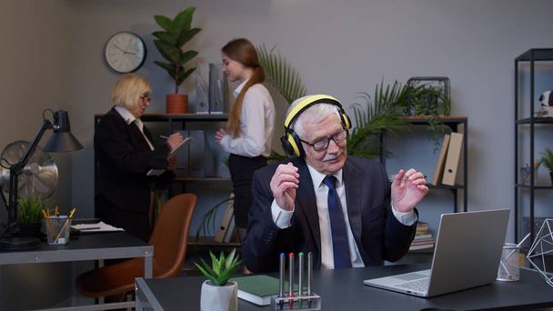 Веселый старший бизнесмен, работающий на портативном ноутбуке, сидит за столом, слушает музыку и танцует - Фото, изображение
