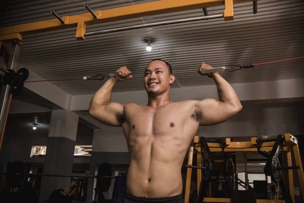 Egy nagy, izmos, félmeztelen ázsiai fickó dupla bicepsz kábeles fürtöket csinál egy kemény edzőteremben. Bicepsz intenzív edzés. - Fotó, kép