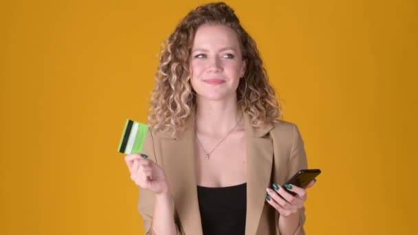 Cheerful expressive young girl with curly hair Using mobile cell phone hold credit bank card makes purchases online shopping isolated on yellow background. People lifestyle concept. - Filmmaterial, Video