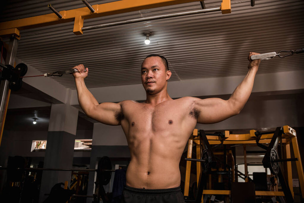 Великий і м'язистий азіатський хлопець стоїть подвійний біцепс кабельних завитків в хардкор спортзалі. Інтенсивно розробляти біцепс
. - Фото, зображення