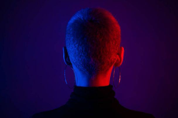 Beautiful young woman with short blonde haircut in neon light stands with her back. - Foto, Imagen