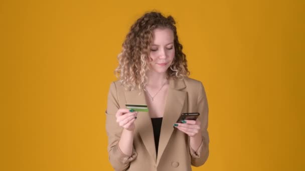 Cheerful expressive young girl with curly hair Using mobile cell phone hold credit bank card makes purchases online shopping isolated on yellow background. People lifestyle concept. - Materiaali, video