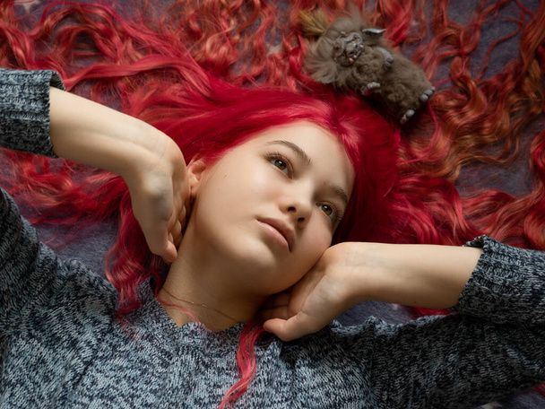 A girl with pink hair in a colorful sweater, lies on a blue blanket, hands hold her neck, on her hair lies a handmade goblin toy - Foto, Imagem