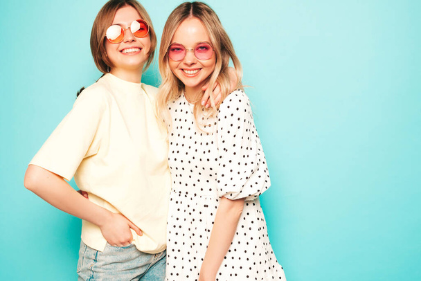 Kaksi nuorta kaunista blondia hymyilevää hipsterinaista trendikkäissä kesävaatteissa. Seksikäs huoleton naiset poseeraa lähellä sinistä seinää studiossa. Trendikäs ja positiivisia malleja hauskaa aurinkolasit - Valokuva, kuva