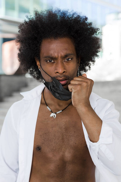 Retrato vertical de hombre negro con pelo afro. Se quita la mascarilla con un gesto serio y desafiante.. - Foto, Imagen