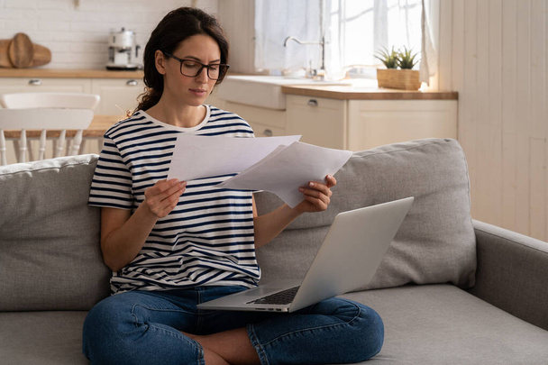 Concentrated millennial woman work at documents sit on couch with laptop, read business paper bills - Photo, Image