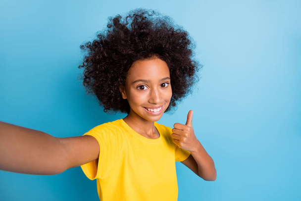 Photo of brunette little afro american girl wear yellow t-shirt take selfie make thumb up isolated on blue color background - Foto, afbeelding