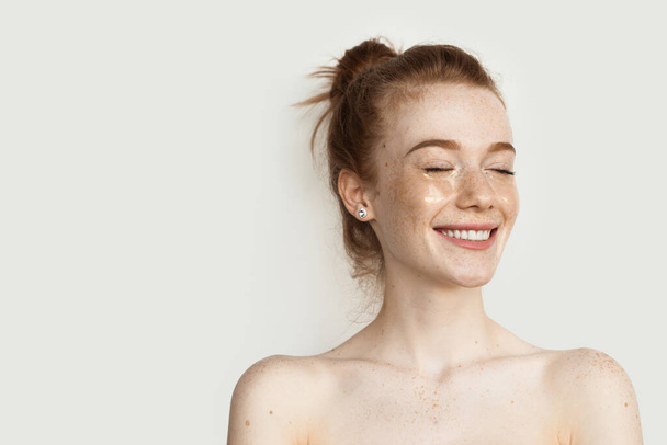 Ginger γυναίκα με μπαλώματα ματιών χαμογελά σε έναν τοίχο στούντιο με γυμνούς ώμους - Φωτογραφία, εικόνα