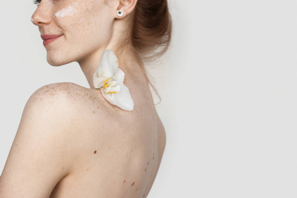 Běloška, která si nanáší krém na tváře, se usmívá, drží na prázdných ramenou květinu - Fotografie, Obrázek