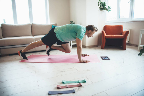 Blond man is met behulp van een tablet tijdens het doen van fitness-oefeningen thuis op een yoga mat - Foto, afbeelding
