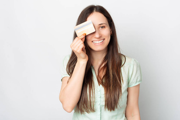Eine nette junge Frau lächelt in die Kamera und hält eine Kreditkarte vor ihrem Auge . - Foto, Bild