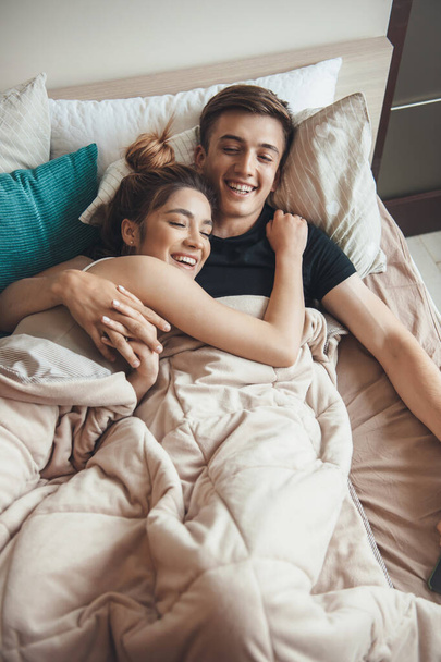 Φωτογραφία πάνω όψης ενός καυκάσιου ζευγαριού ξαπλωμένου στο κρεβάτι καλυμμένου με μια κουβέρτα που χαμογελά και αγκαλιάζει - Φωτογραφία, εικόνα