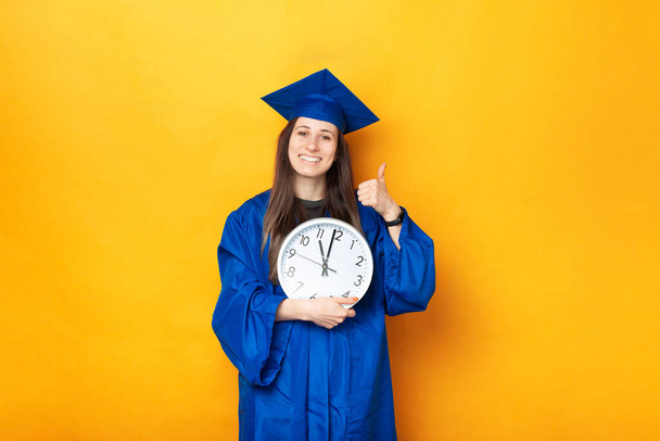 Una bonita foto de un estudiante alegre sosteniendo un gran reloj blanco vestido con un uniforme de graduación cerca de una pared amarilla . - Foto, imagen