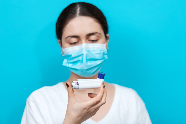 Bronchial asthma. Blurry portrait of a woman in a medical mask holding an inhaler in front of her and looking it. Blue background. Copy space. - Photo, Image