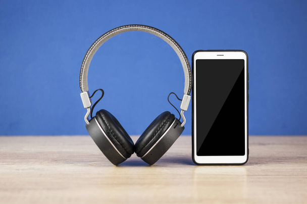 Černá sluchátka a chytrý telefon na dřevěném stole proti modré tapety pozadí - Fotografie, Obrázek