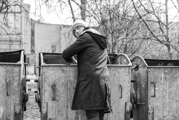 monochrome photo of homeless young guy rummages in trash cans in the park. the problem of homeless and unemployed emigrants. Lifestyle of tramp, living in the streets - Photo, Image
