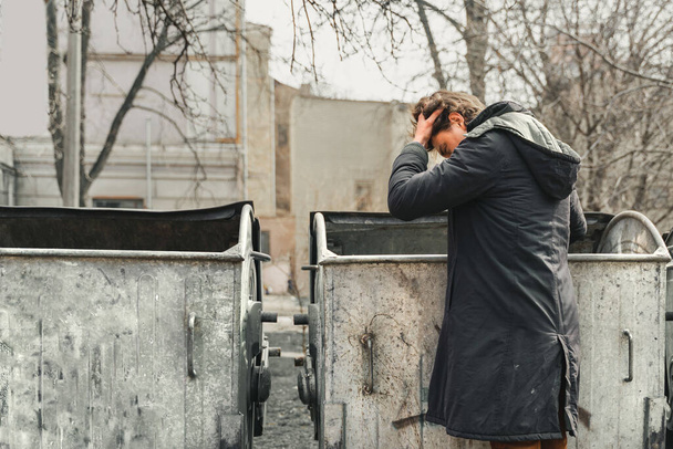 zdesperowany młodzieniec trzymający głowę, szukający jedzenia w śmietnikach. Samotny człowiek, który stracił pracę szukając jedzenia w śmietniku. problem osób bezdomnych i bezrobotnych. Styl życia włóczęgi, żyjącej na ulicy - Zdjęcie, obraz