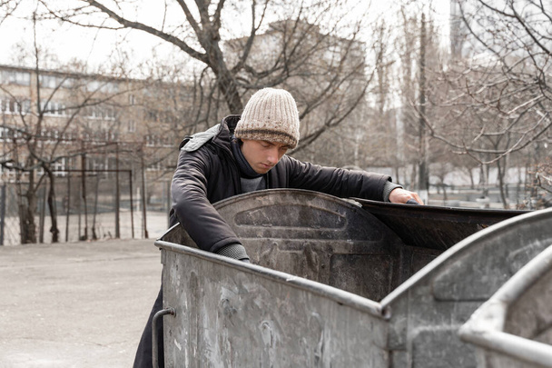 un pobre caucásico está buscando comida y objetos de valor en un cubo de basura en la calle. el problema de las personas sin hogar y desempleadas - Foto, Imagen