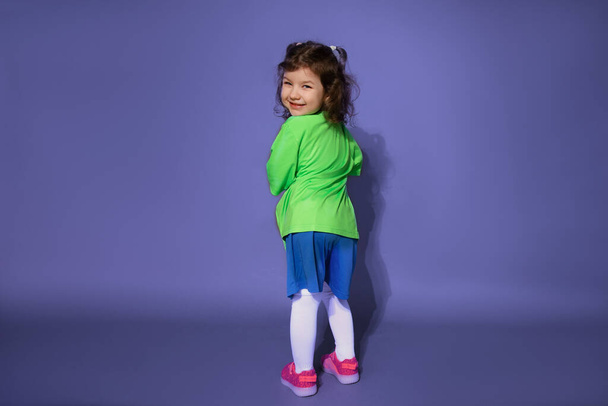 Забавная маленькая девочка в футбольной форме стоит спиной на фиолетовом фоне и смотрит в рамку - Фото, изображение