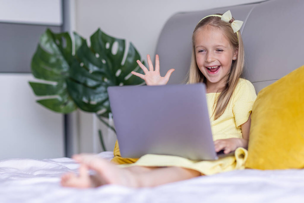 Cute little caucasian girl with blonde hair in fashionable dress illuminating yellow color sitting at home during coronavirus pandemic quarantine and using laptop. Stay at home during covid-19 - Fotografie, Obrázek
