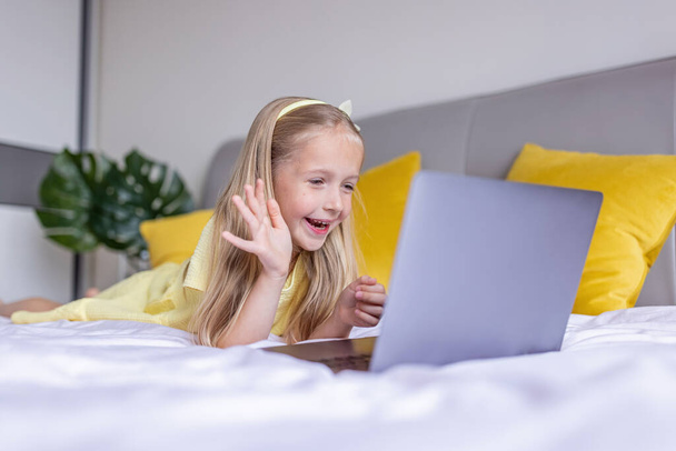 Cute little caucasian girl with blonde hair in fashionable dress illuminating yellow color sitting at home during coronavirus pandemic quarantine and using laptop. Stay at home during covid-19 - Foto, Imagem