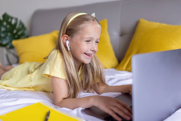 Linda niña caucásica con cabello rubio en vestido de moda iluminando el color amarillo sentado en casa durante la cuarentena pandémica coronavirus y el uso de la computadora portátil. Permanecer en casa durante covid-19 - Foto, imagen