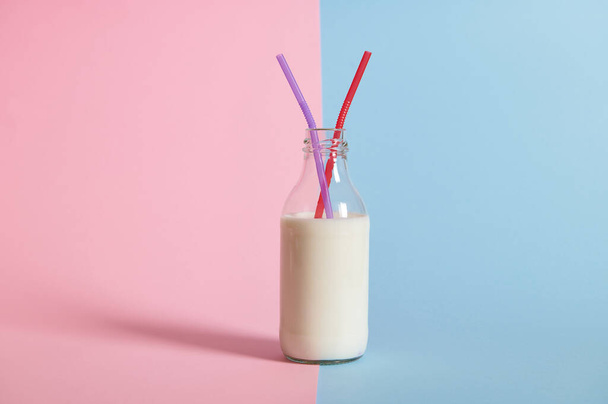 Primer plano de botella transparente con leche y dos pajitas en ella, aislada sobre fondo bicolor azul y rosa con espacio para texto - Foto, Imagen