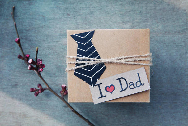 Handmade craft box gifts with paper tie, text I love dad, branche, flowers on blue wooden background. Concept International Father's day, birthday, eco. Minimalism. Top view, copy space - 写真・画像