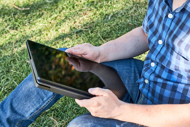 Jeune homme méconnaissable utilisant une tablette à l'extérieur, assis sur l'herbe. Concepts du télétravail et de l'étude. - Photo, image