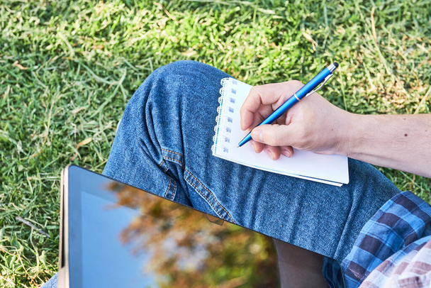 Nahaufnahme, unkenntlicher junger Mann, der mit einem Stift in ein Notizbuch schreibt, im Gras sitzt, mit einem Tablet studiert oder im Freien arbeitet. - Foto, Bild
