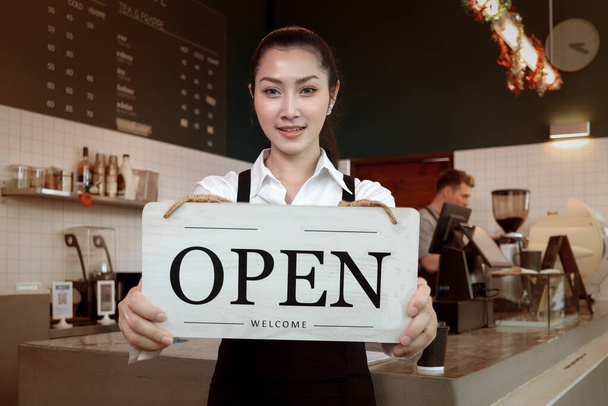 Joven hermosa mujer asiática barista sosteniendo letrero abierto amplio en restaurante y cafetería para decir a los clientes que la tienda está lista para el servicio. - Foto, imagen