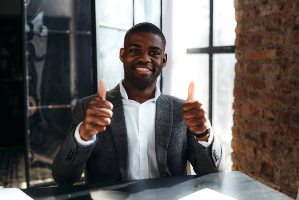 Happy black-skinned businessman in suit sitting at the table smiling at the camera and showing thumbs up - Фото, изображение