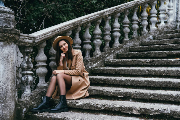 Beautiful young woman wearing hat and jacket, sitting on stairs. Travel concept. - Zdjęcie, obraz