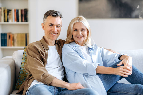 Portrait of mature couple in love. Middle aged joyful caucasian marriage couple spend time together at home, relaxing on a sofa in the living room, dressed in casual stylish clothes, smiling at camera - Fotó, kép