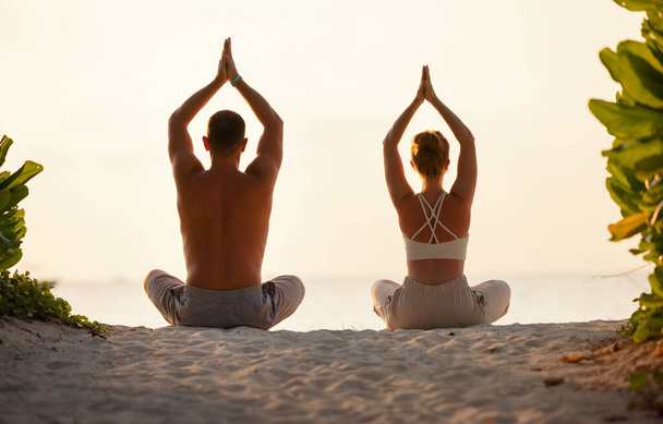 Visão traseira de homem e mulher em roupas esportivas praticando postura de lótus com as mãos Namaste acima das cabeças na costa - Foto, Imagem