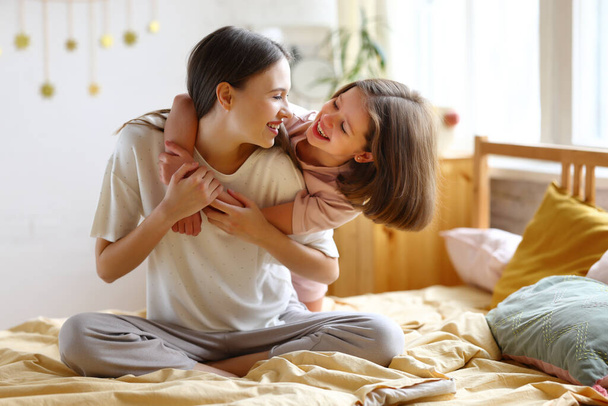 Happy moments. Young loving mother cuddling with adorable little girl daughter while sitting together on bed, looking at each other and smiling. Older and younger sister enjoying carefree time at home - Foto, Bild