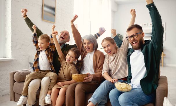Celebrating goal. Excited big multi generation family, grandparents, young mother and father with little kids sitting on coach in living room and watching football match on tv, having fun together - Фото, изображение