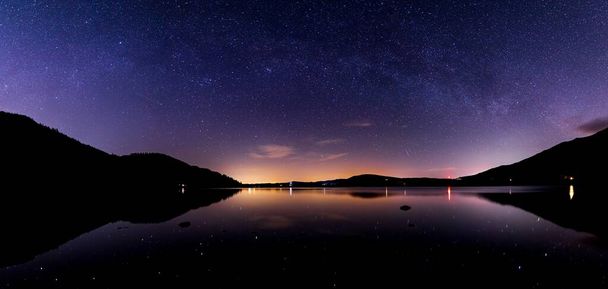 A View of Bassenthwaite lake at night with a faint milky way arching over the lake and stars and lights reflecting on the lake - Photo, Image