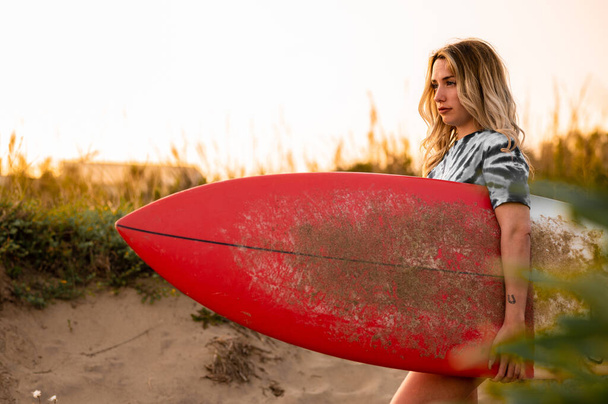 A young blonde female surfer walking with her surfing board at the beach - Photo, image