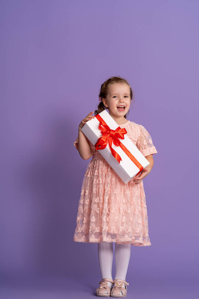 little cute girl with gift box with red ribbon on purple background. birthday of small child. a happy child holds a gift in his hands. a birthday present. concept of celebrating, giving and receiving - Foto, Imagem