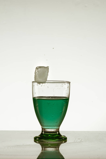 an ice cube falling into a glass of green drink isolated on the reflective surface - Photo, Image