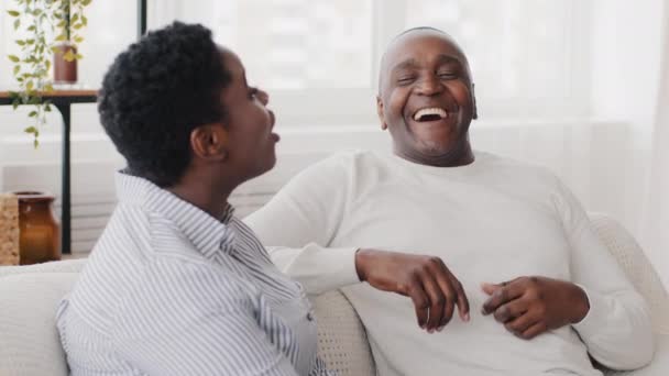 家族のカップルアフロアメリカの女性の娘と黒の大人の中年の男性の父親は、家庭のソファのチャット会話に座っている。室内で笑っている二世代の民族の妻と老人 - 映像、動画