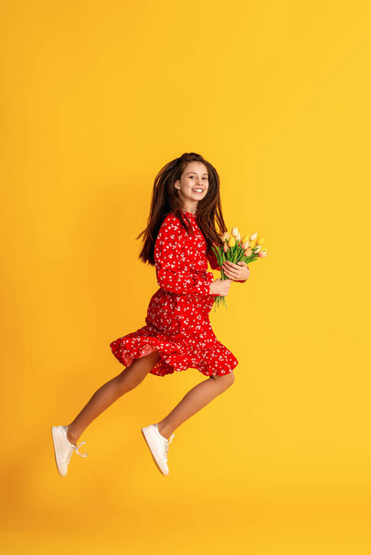 Cute charming dreamy teenage girl, with bouquet tulips, in red dress with floral print, lies on yellow studio background. Caucasian brunette jumping on background colored wall.Copy space - Фото, зображення
