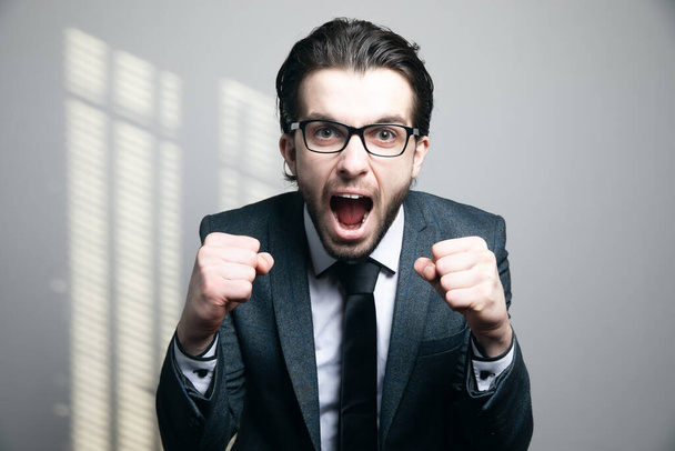 man in een pak en bril schreeuwen gebalde vuisten op een grijze achtergrond - Foto, afbeelding