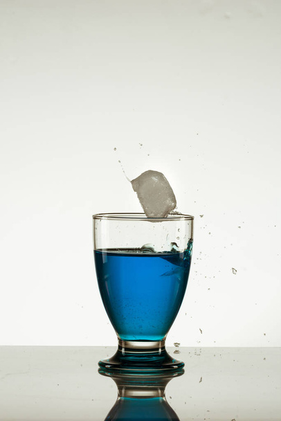 an ice cube falling into a glass of blue drink isolated on the reflective surface - Фото, изображение