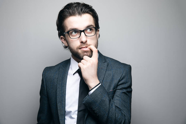 man in a suit and glasses bites his finger on a gray background - Photo, Image