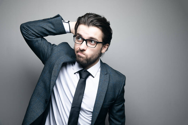 a man in a suit and glasses looks thoughtfully scratches his head on a gray background - Foto, Imagem
