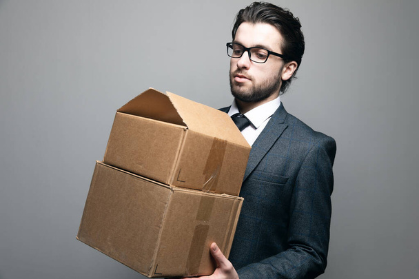 een man in pak en bril staat met dozen in zijn handen op een grijze achtergrond - Foto, afbeelding